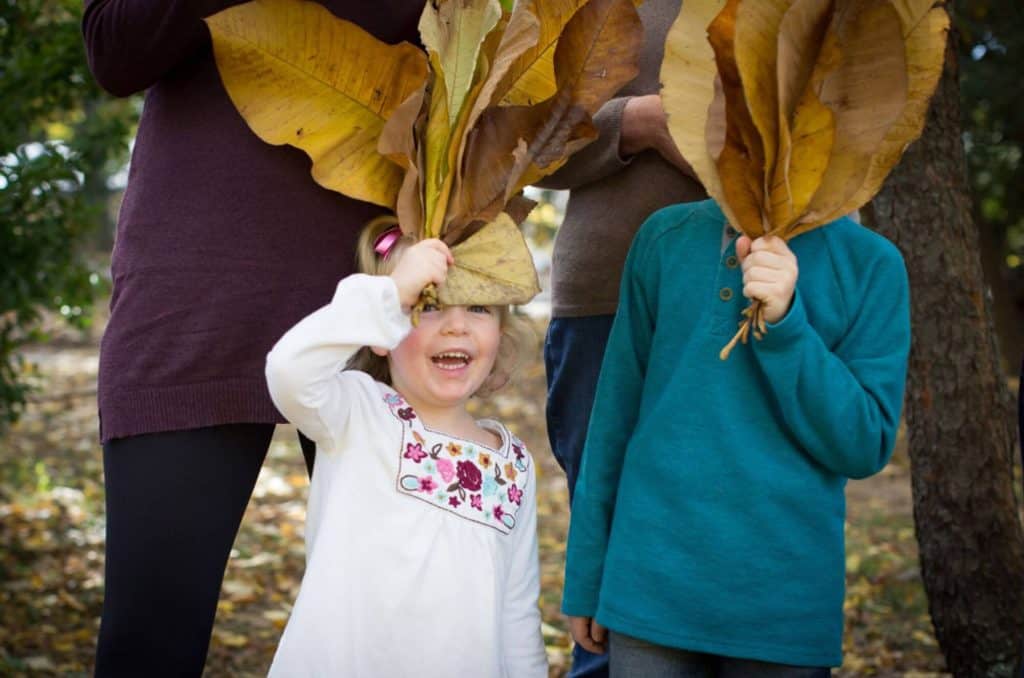 a family poses with leaves covering their face and the girl in the dress peeks out and laughs