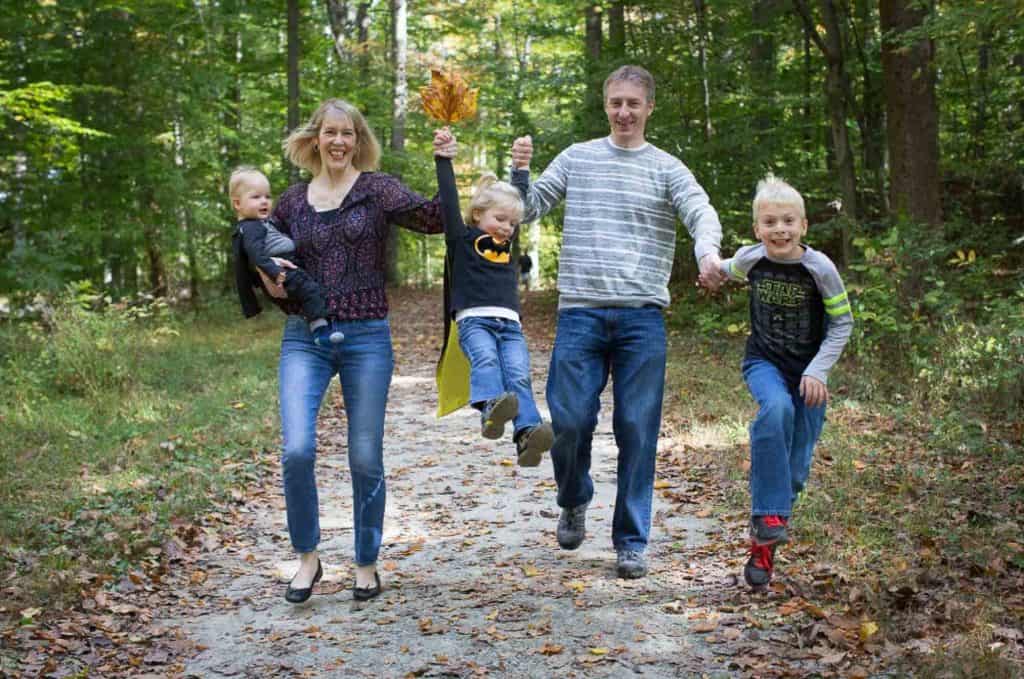 a family runs down a fall path, the girl holds leaves and is wearing a Batman cape like the baby, the oldest son wears a Star Wars tee shirt