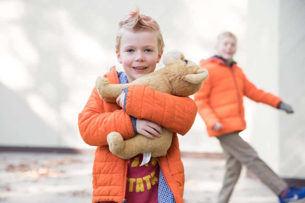 a girl poses with her short hair and puffy orange jacket, her brother dances through the background