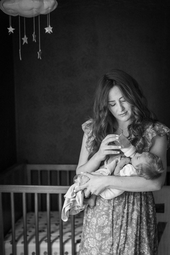 a mom feeds her newborn a bottle as she stands near his crib