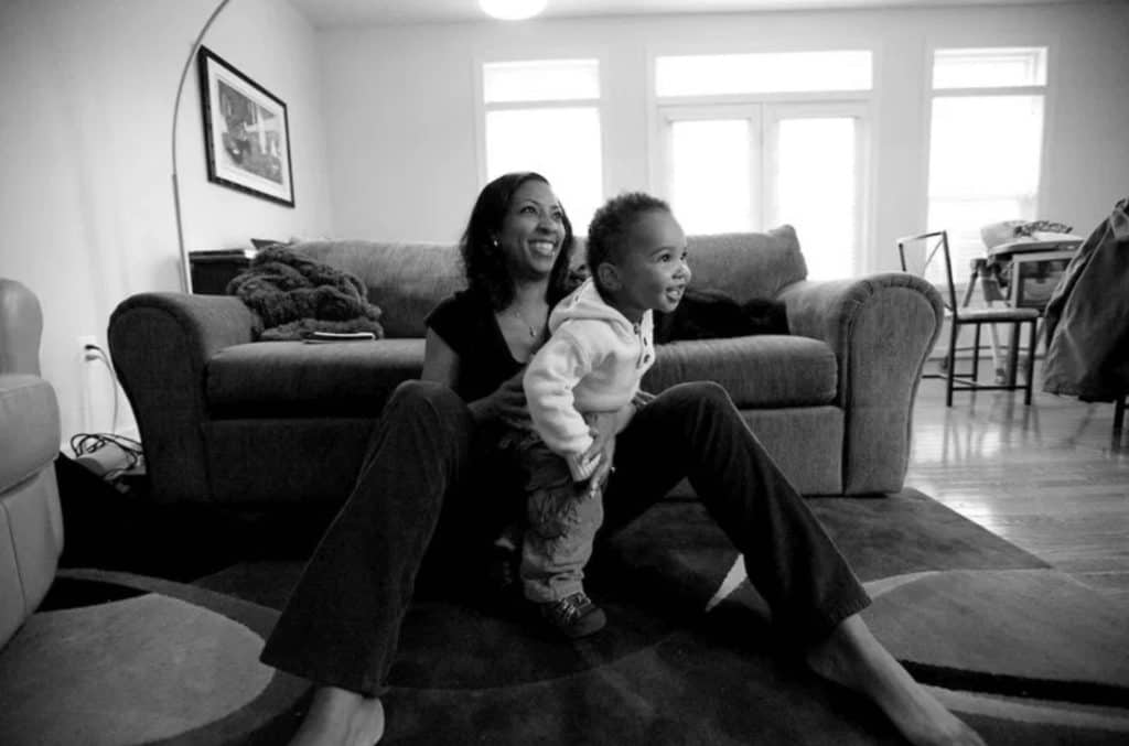 a mom sits beneath the couch, tickling her toddler and smiling towards her partner