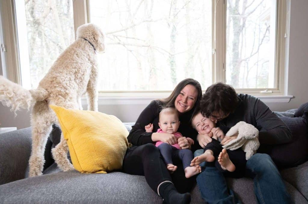 parents cuddle their children on the couch while the dog looks out the window at the world behind them