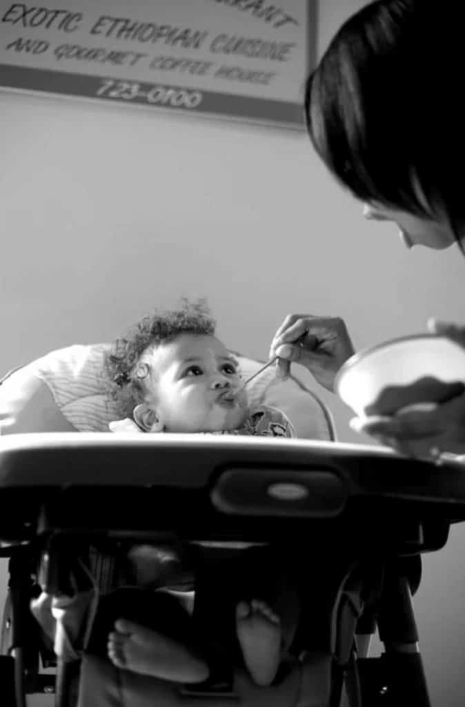 a mom spoons solid foods into her baby's mouth as he sits in his high chair, her mouth is open as if to say, "open wide!"